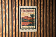 A metal sign with a vintage-style illustration of a palm tree and a landscape, mounted on a wooden wall.