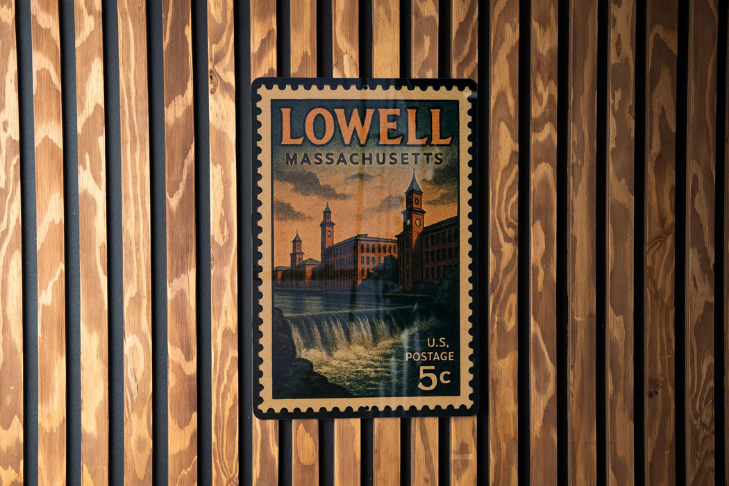 A vintage postage stamp from Lowell, Massachusetts, featuring a building and a waterfall, is displayed on a wooden wall.