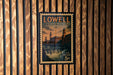 A vintage postage stamp from Lowell, Massachusetts, featuring a building and a waterfall, is displayed on a wooden wall.