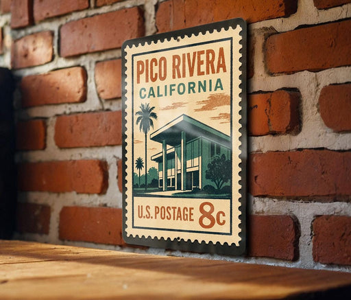 A vintage-style postage stamp from Pico Rivera, California, featuring a building and palm trees, is displayed on a brick wall.