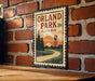 A vintage-style sign for Orland Park, Illinois, featuring a scenic illustration of a winding road, pine trees, and a water tower against a sunset backdrop.