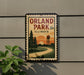 A vintage-style sign for Orland Park, Illinois, featuring a scenic landscape with a winding road and a water tower in the background.
