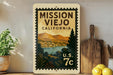 A vintage-style metal sign with a postage stamp design, featuring a scenic landscape of mountains, a river, and a town, mounted on a wall next to a wooden cutting board and some potted plants.