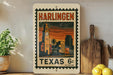 A vintage postage stamp from Harlingen, Texas, featuring a cityscape with a clock tower, is displayed on a shelf next to a wooden cutting board and some lemons.