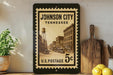 A vintage-style postage stamp from Johnson City, Tennessee, featuring an old street scene and a water tower, displayed on a shelf next to a wooden cutting board and some lemons.