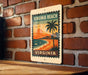 A vintage-style metal sign with a tropical beach scene and palm trees, mounted on a brick wall.