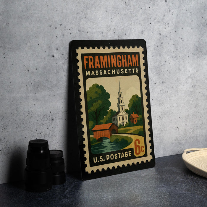 A framed postage stamp from Framingham, Massachusetts, depicting a church and a bridge, placed on a countertop next to some camera equipment.
