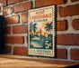 A vintage-style postage stamp from the United States, featuring a tropical beach scene with palm trees and a boat, mounted on a brick wall.