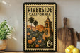 A vintage-style postage stamp from Riverside, California, featuring a bell tower and palm trees, is displayed on a shelf next to a wooden cutting board and a potted plant.