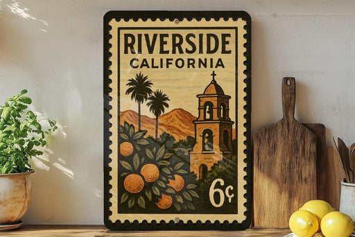 A vintage-style postage stamp from Riverside, California, featuring a bell tower and palm trees, is displayed on a shelf next to a wooden cutting board and a potted plant.