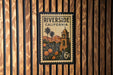 A vintage postage stamp from Riverside, California, featuring a bell tower and palm trees, mounted on a wooden wall.
