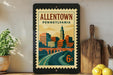 a framed poster of the town of Allentown, Pennsylvania, with a cityscape and a bridge depicted on it. The poster is placed on a shelf next to a wooden cutting board and a potted plant.