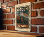 A vintage-style metal sign with the text "OGDEN UTAH" is displayed on a brick wall, featuring an illustration of a cityscape with mountains in the background.