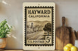 A vintage-style stamp from the United States Postal Service, featuring an illustration of a town in California with a road and buildings. The stamp is mounted on a wall, and there are some kitchen utensils and a potted plant visible in the foreground.
