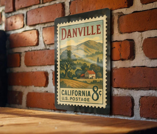 A vintage-style metal postage stamp from California, featuring a rural landscape and a house, is displayed on a brick wall.