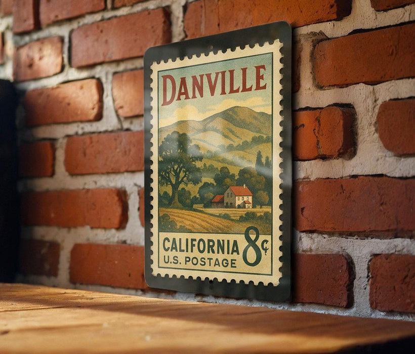 A vintage-style metal postage stamp from California, featuring a rural landscape and a house, is displayed on a brick wall.