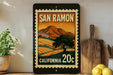 A framed poster of a scenic landscape with mountains and a tree, featuring the text "SAN RAMON" and "CALIFORNIA 20c" on it, is displayed on a shelf next to a wooden cutting board and some potted plants.