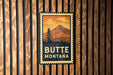 A vintage-style metal sign with the text "BUTTE MONTANA" displayed prominently against a wooden background.