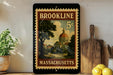 A vintage-style postage stamp from Massachusetts featuring a building and trees, placed on a wooden surface next to a wooden cutting board and a potted plant.