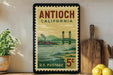 A vintage postage stamp from California, featuring a bridge and a landscape, is displayed on a shelf next to a wooden cutting board and some lemons.