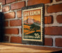 A metal sign with a vintage-style illustration of a landscape, mounted on a brick wall.