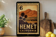 A framed stamp-like artwork depicting a scenic landscape with palm trees and mountains, hanging on a wall next to a wooden cutting board and a potted plant.