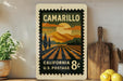 A vintage postage stamp from California, featuring a landscape with a sun and mountains, is displayed on a shelf next to a wooden cutting board and some potted plants.