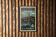 A vintage postage stamp from New York featuring a boat on a river, mounted on a wooden wall.