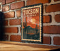 A vintage-style poster of the city of Tucson, Arizona, featuring a desert landscape with cacti and mountains in the background, mounted on a brick wall.