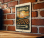 A vintage postage stamp from California, featuring a landscape with a sunset and a field, is displayed on a brick wall.
