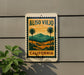 A vintage-style sign with a palm tree and landscape design, mounted on a wall, with a plant to the left.
