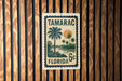 A vintage-style stamp or sign with the text "TAMARAC FLORIDA 6c" printed on it, featuring a palm tree and a sunset landscape.
