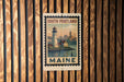 A metal sign with a lighthouse and a cityscape, mounted on a wooden wall.
