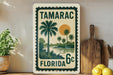 A vintage-style stamp depicting a tropical landscape with palm trees, a sun, and a body of water, with the text "TAMARAC FLORIDA 6c".