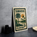 A metal sign with a tropical landscape and palm trees, featuring the text "TAMARAC FLORIDA 6c" on it, is displayed on a table next to some black camera lenses.