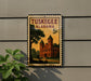 A postage stamp from Alabama featuring a building with a tower, mounted on a wall next to a plant.