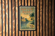 A vintage-style postage stamp from the United States, featuring a tropical beach scene with palm trees and a house, mounted on a wooden wall.