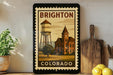 A framed poster of a water tower and a building, with the text "BRIGHTON COLORADO" on it, is displayed on a wall next to a wooden cutting board and a potted plant.