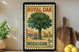 A vintage-style postage stamp featuring a tree and a store, with the text "ROYAL OAK MICHIGAN 5¢ U.S. POSTAGE" printed on it. The stamp is displayed on a wall, with a wooden cutting board and a potted plant visible in the foreground.