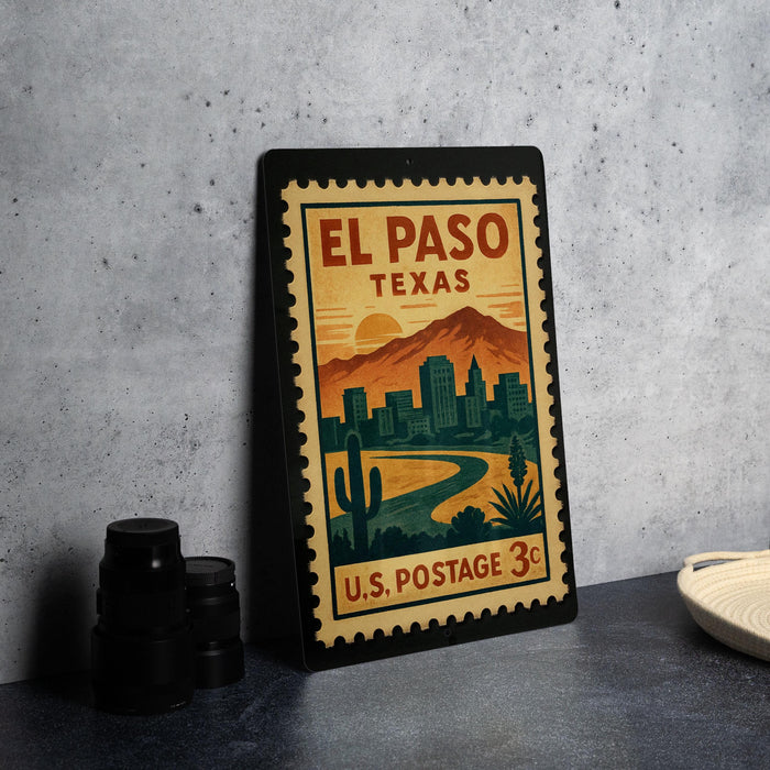 A vintage-style postage stamp from El Paso, Texas, featuring a desert landscape with mountains and buildings, placed on a countertop next to some black camera lenses.