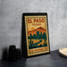 A vintage-style postage stamp from El Paso, Texas, featuring a desert landscape with mountains and buildings, placed on a countertop next to some black camera lenses.
