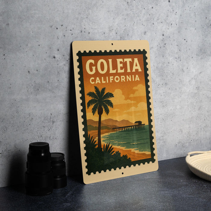 A vintage-style metal sign with the text "GOLETA CALIFORNIA" and an illustration of a palm tree and a pier, placed on a gray countertop next to some black camera lenses.