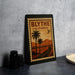 A framed poster of a road leading to palm trees, with a vintage postage stamp design, is displayed on a table.