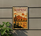 A vintage-style metal sign with a desert landscape and mountains, featuring a cactus and a sun, mounted on a wall.