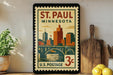 A framed postage stamp from St. Paul, Minnesota, depicting a cityscape with a bridge and a river, is displayed on a shelf next to a wooden cutting board and some lemons.