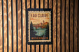A vintage-style poster of Eau Claire, Wisconsin, featuring a bridge and a river, is displayed on a wooden wall.