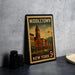 A vintage-style metal stamp depicting a town scene with a clock tower, surrounded by buildings and a street lamp, placed on a countertop next to some black camera lenses.