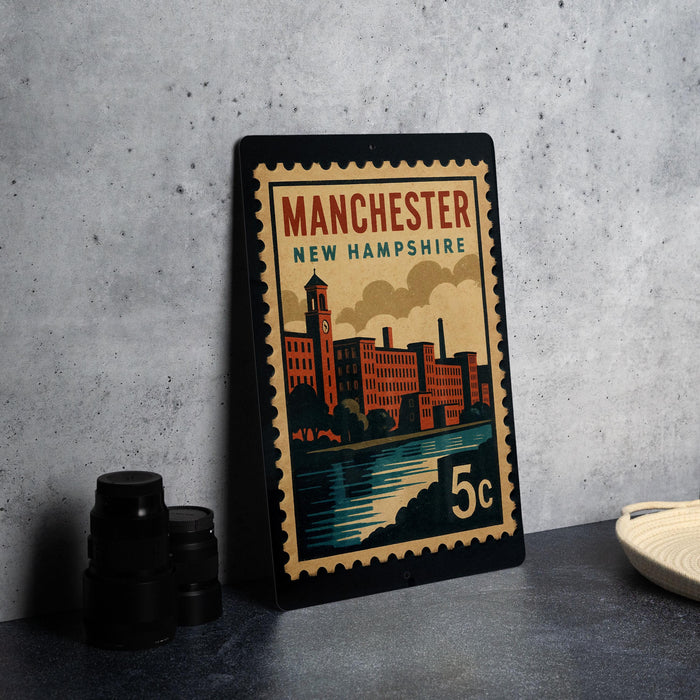 A vintage-style metal stamp depicting the city of Manchester, New Hampshire, with a building and river in the background.