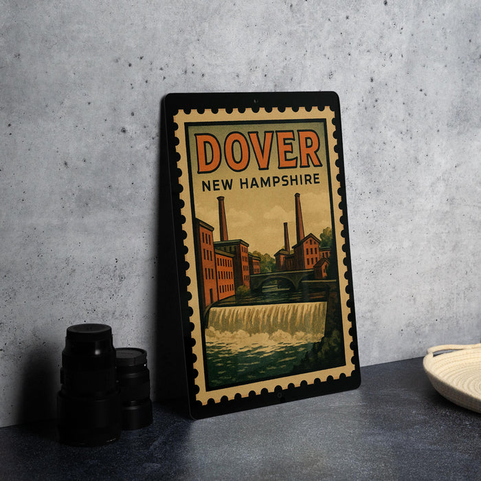A vintage-style metal poster or sign with the text "DOVER NEW HAMPSHIRE" and an illustration of a waterfall and buildings, placed on a countertop next to some black camera lenses.