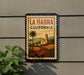A framed poster of La Habra, California, featuring a scenic illustration of a road, palm trees, and a church.